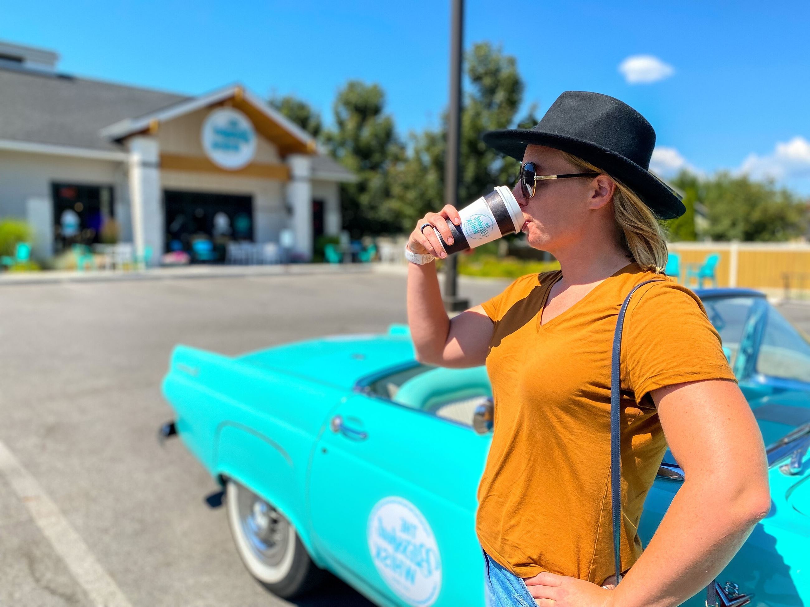Owner standing in front of blue car sipping coffee in parking lot of new business.